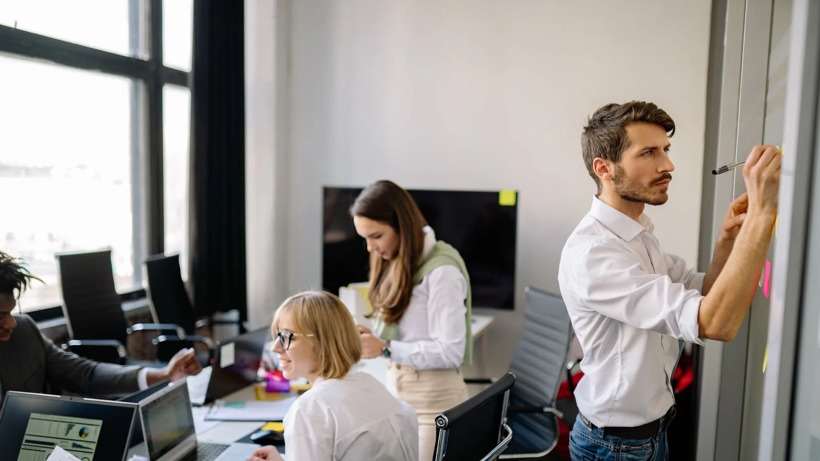 Team in a bright office: a man posts sticky notes on a wall while colleagues work at laptops