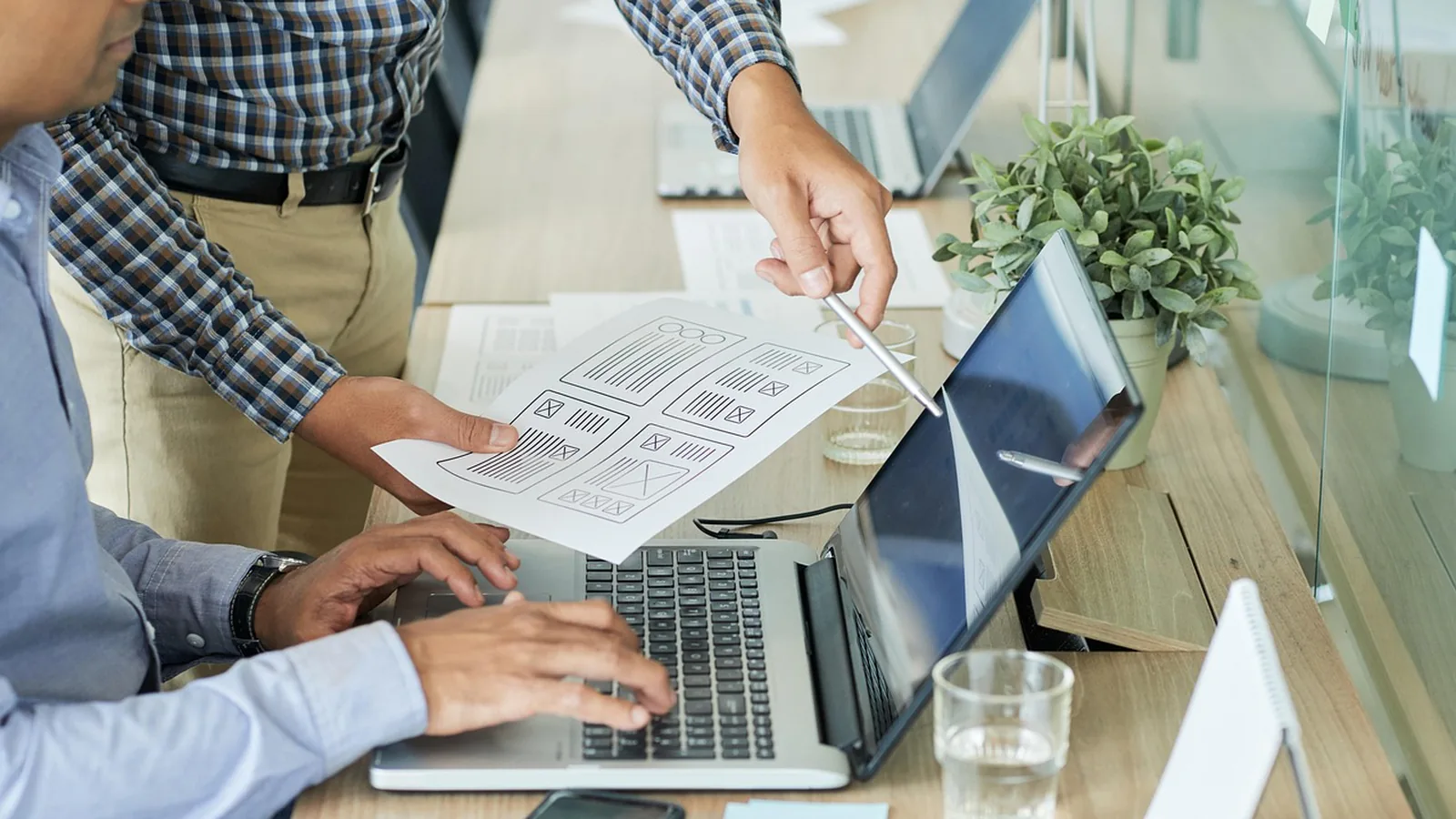 Two colleagues review a printed website wireframe beside an open laptop on a shared desk