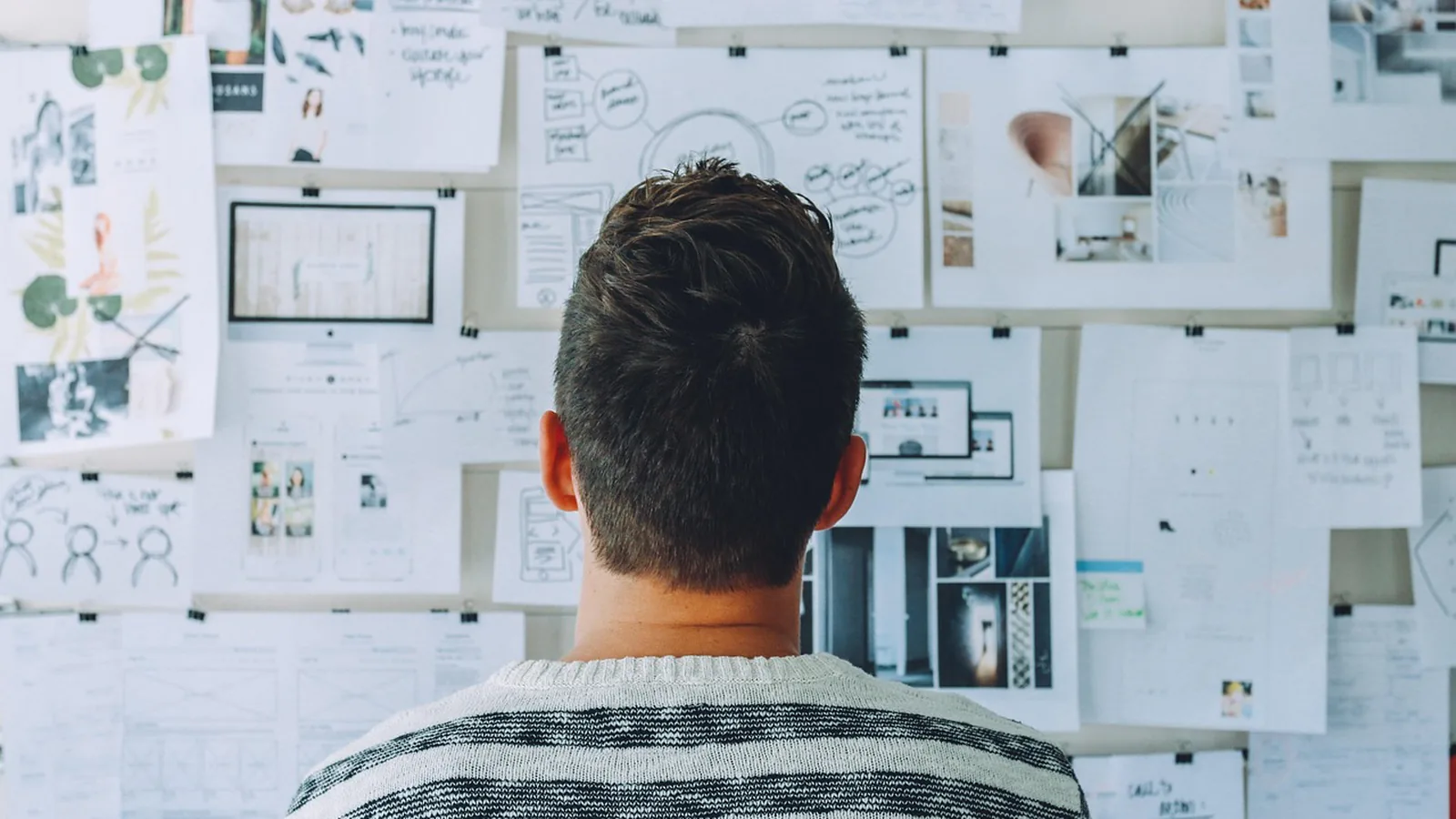 Man seen from behind studying a wall of pinned wireframes and sketches while planning site structure