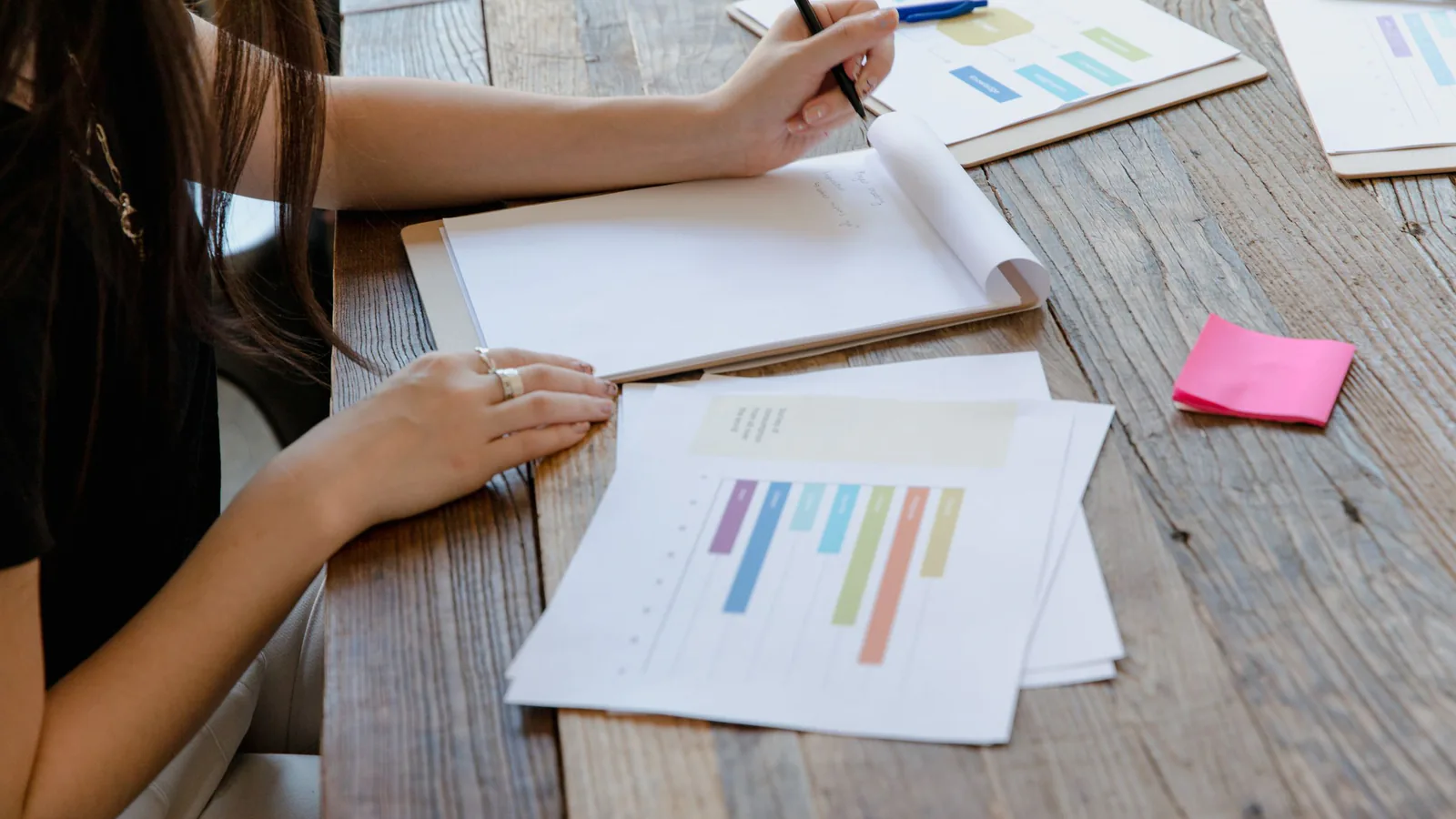 Person writing notes beside printed bar charts on a wooden table, as when logging AI citation results