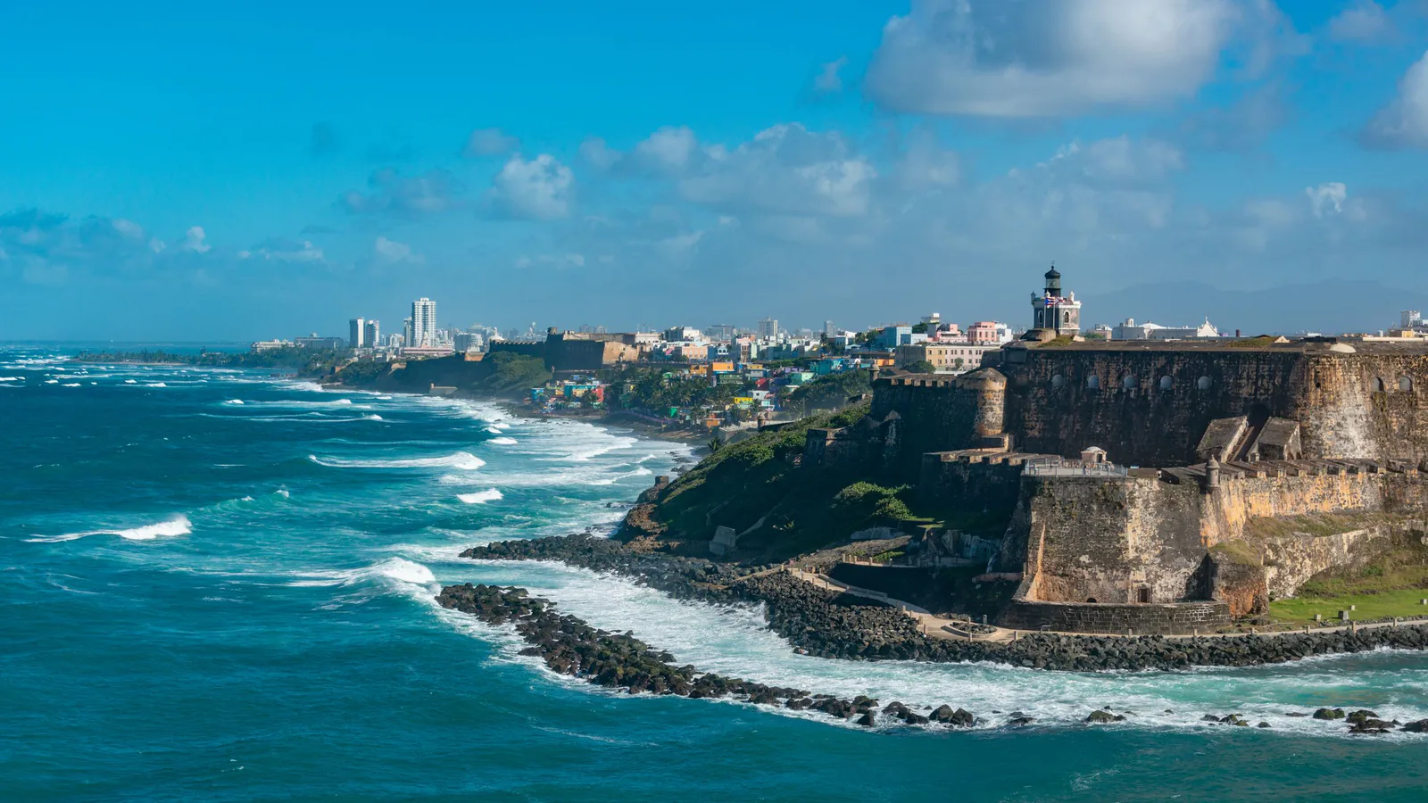 El Morro fortress and lighthouse above rough turquoise surf, with the San Juan skyline beyond