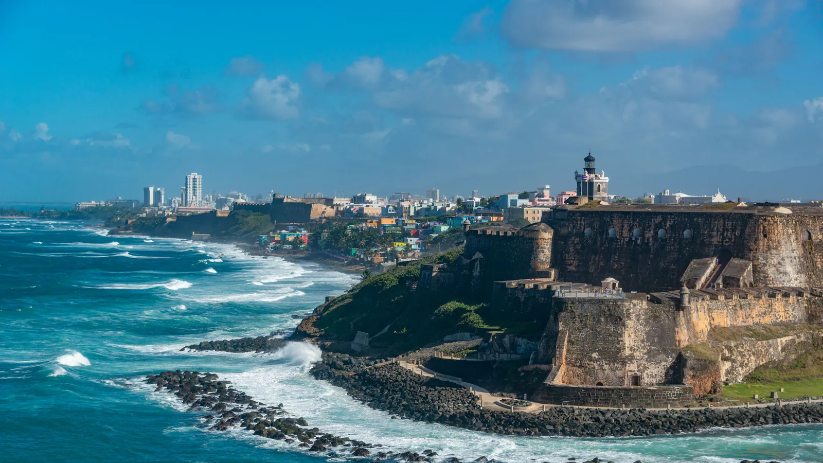 Stone fortress and lighthouse on a rocky San Juan headland, waves breaking below, city skyline in the distance