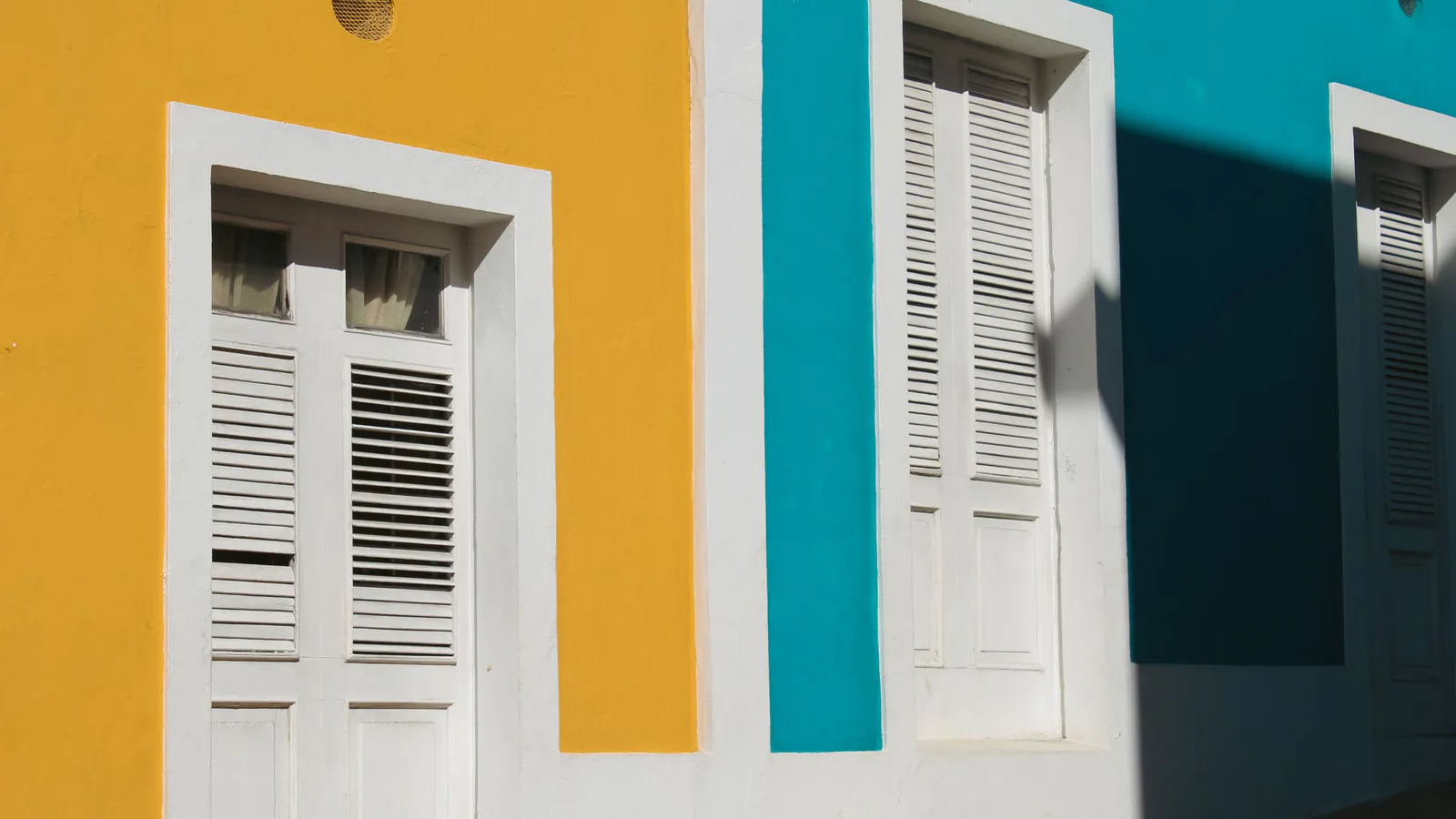 Yellow and teal painted walls with white shuttered doors in bright sun, suggesting a place of business