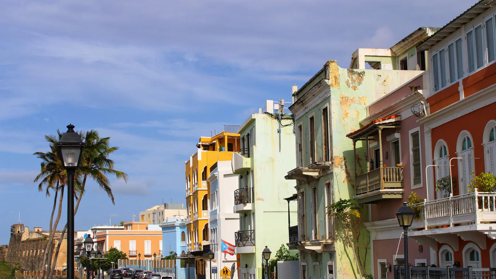 Pastel and yellow townhouses on a sunlit Old San Juan street, with palm trees and a stone fortress wall