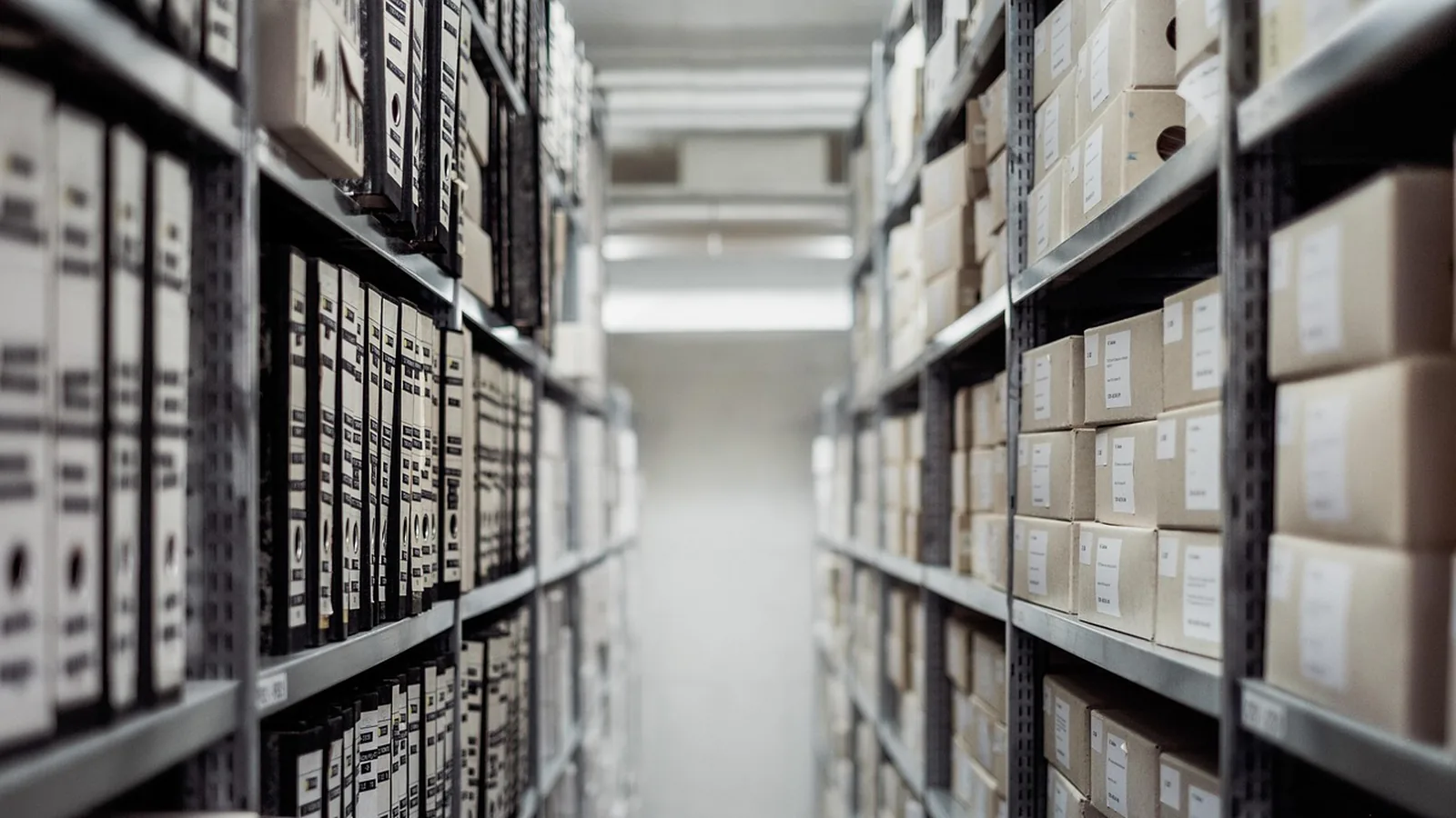 Rows of archive boxes and binders on metal shelves along a narrow aisle, suggesting records kept in order