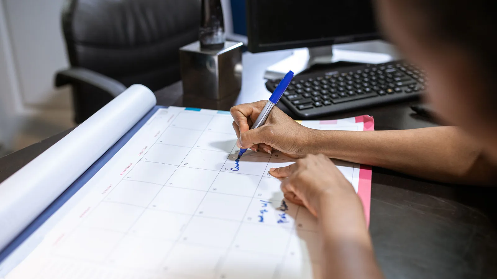 Person writing in blue pen on a monthly desk calendar planner beside a keyboard and monitor