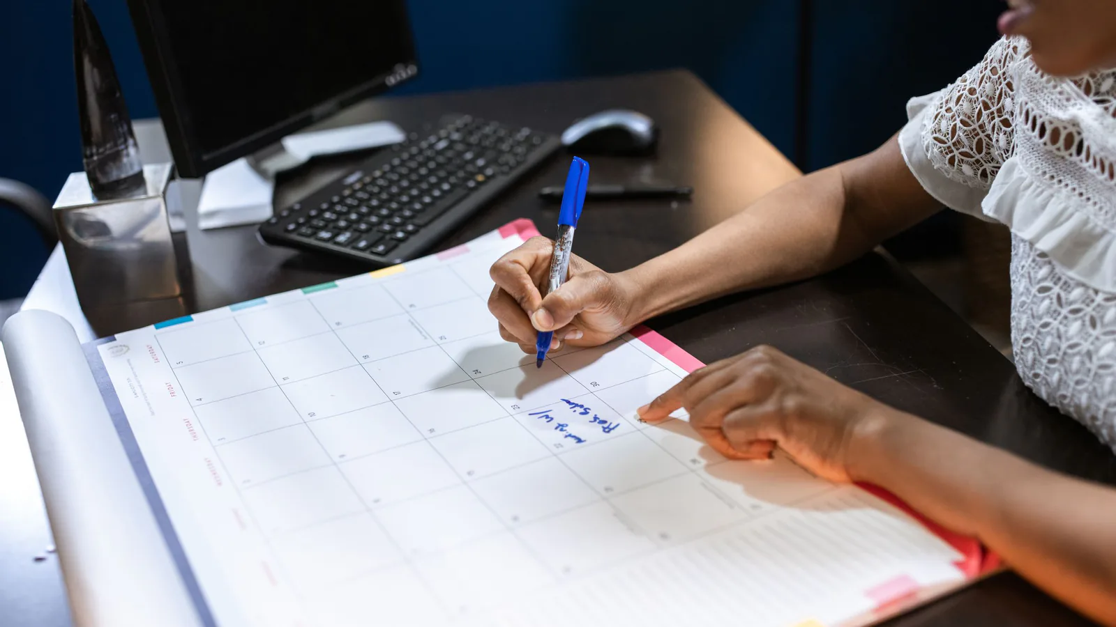 Person writing with a blue marker on a large monthly calendar planner laid on a dark desk