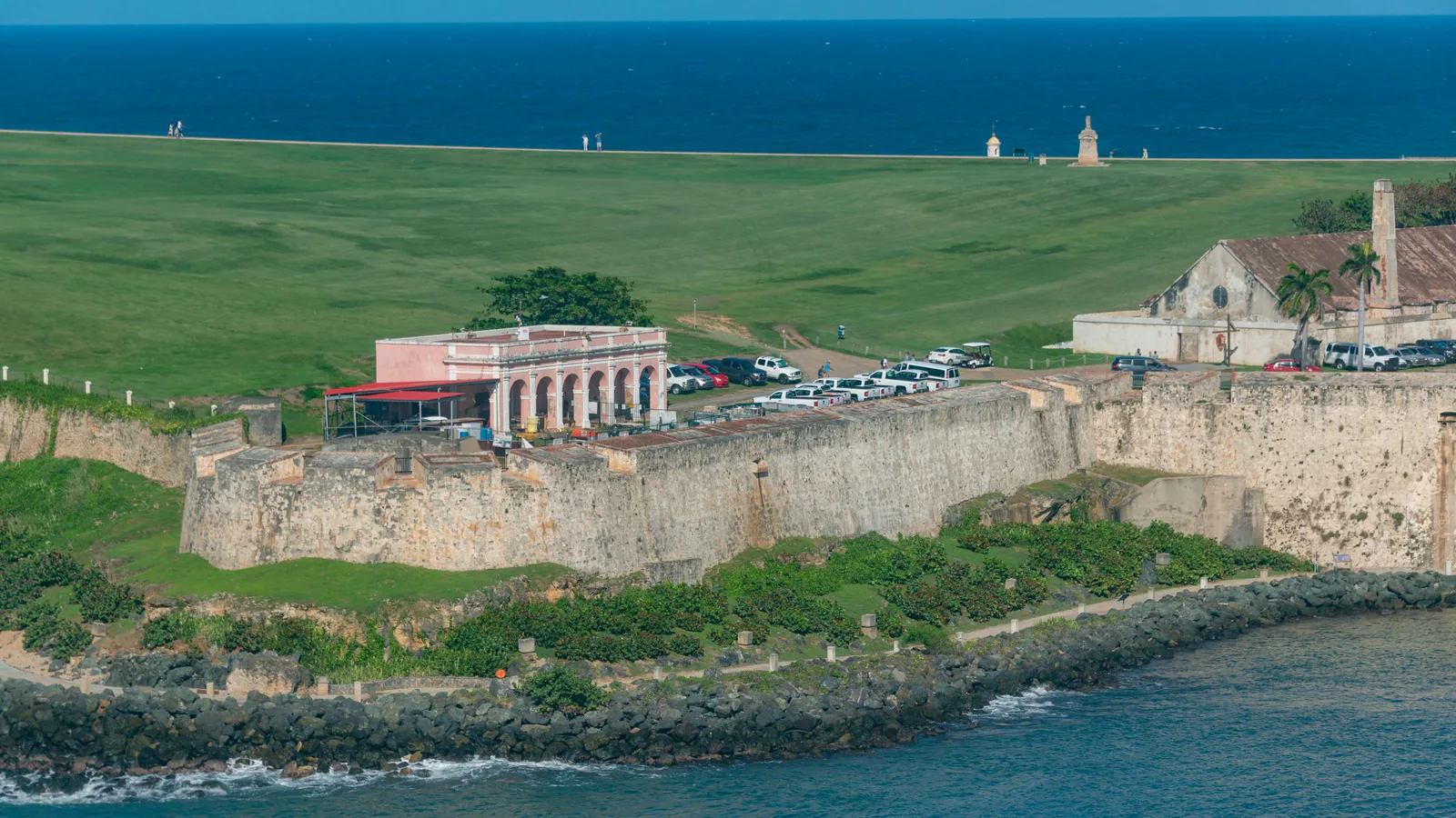 Aerial view of an old stone fortress wall above a rocky shoreline, with a green lawn and the sea beyond
