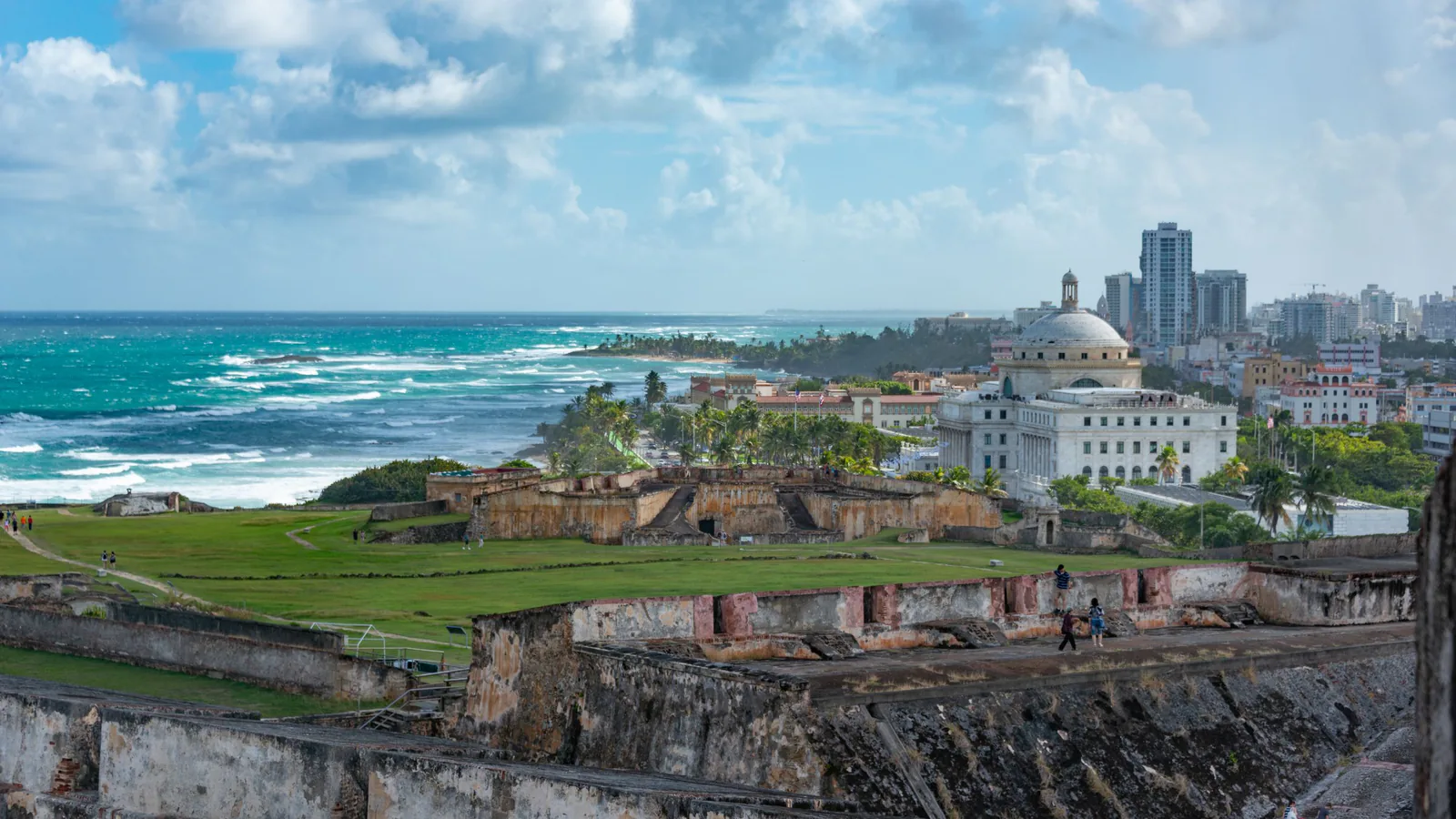 Old stone fortress walls above a turquoise coastline, with the San Juan skyline and domed Capitol in Puerto Rico