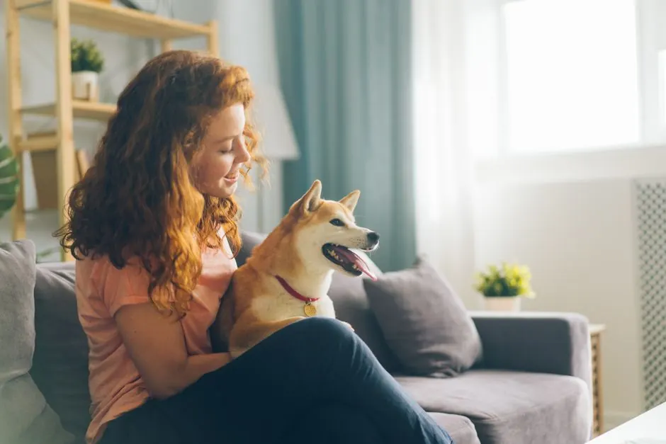 Una mujer pelirroja en un sofá con un shiba inu con collar rojo sentado en su regazo.