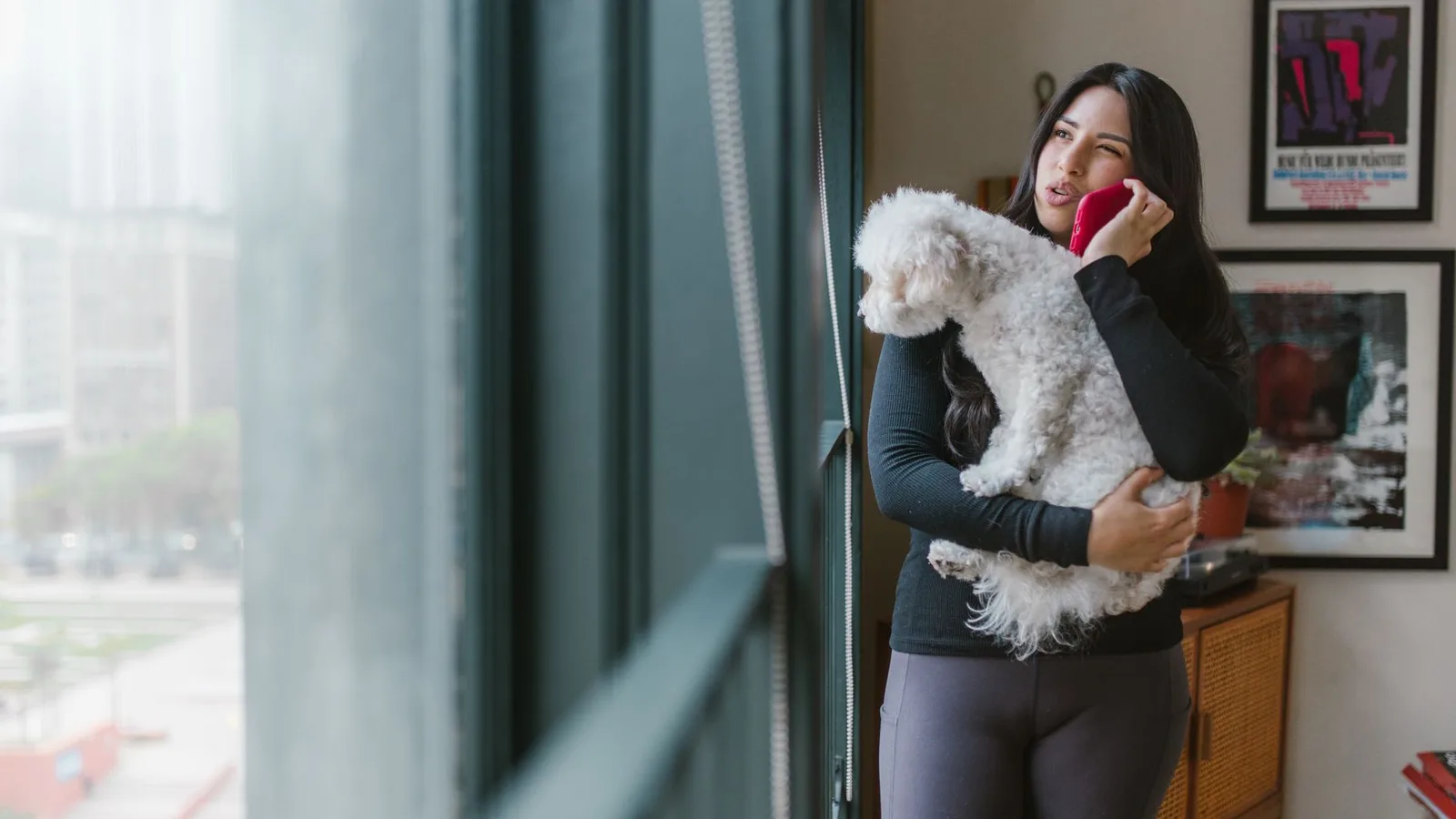 A woman holds a small white dog in one arm and a phone to her ear by a window.