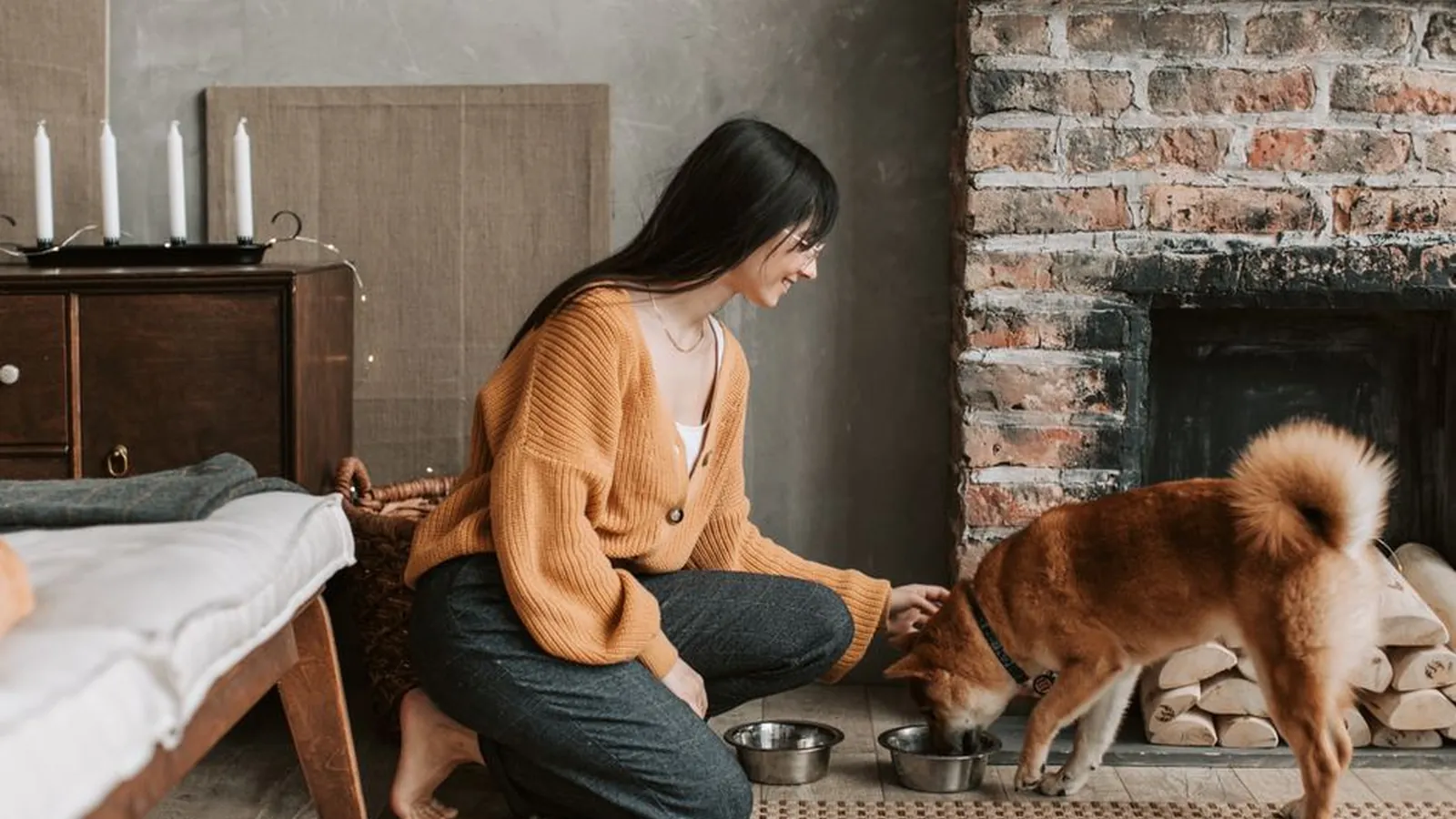 A woman kneeling on the floor of a living room smiles as a Shiba Inu eats from a steel bowl beside a brick fireplace.