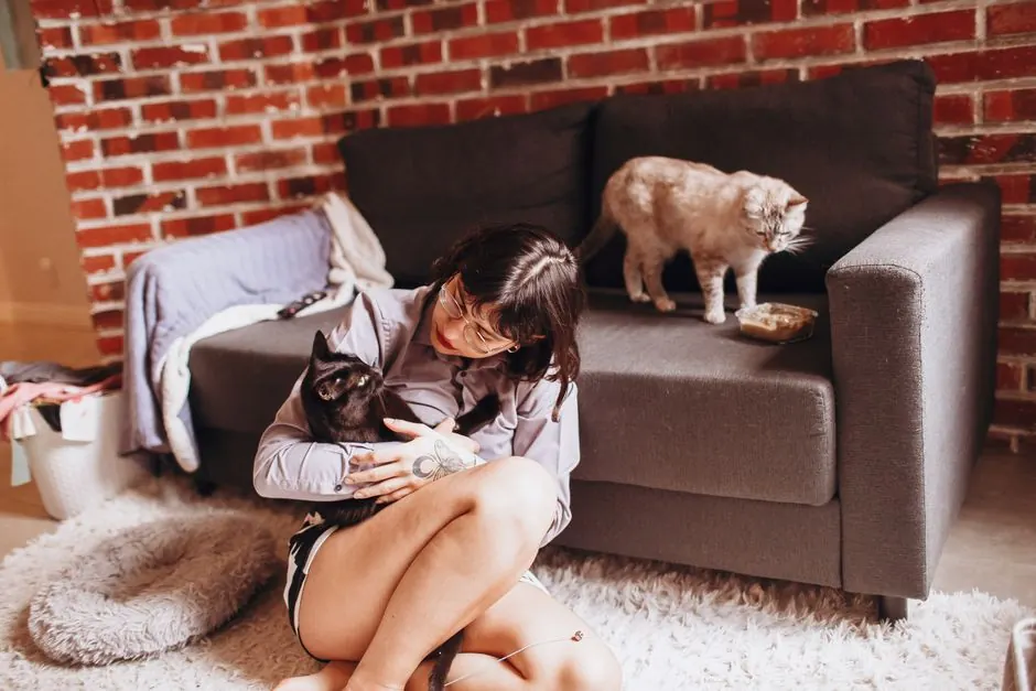 A woman sits on a rug holding a black cat while a second cat stands on the sofa beside a small food dish.
