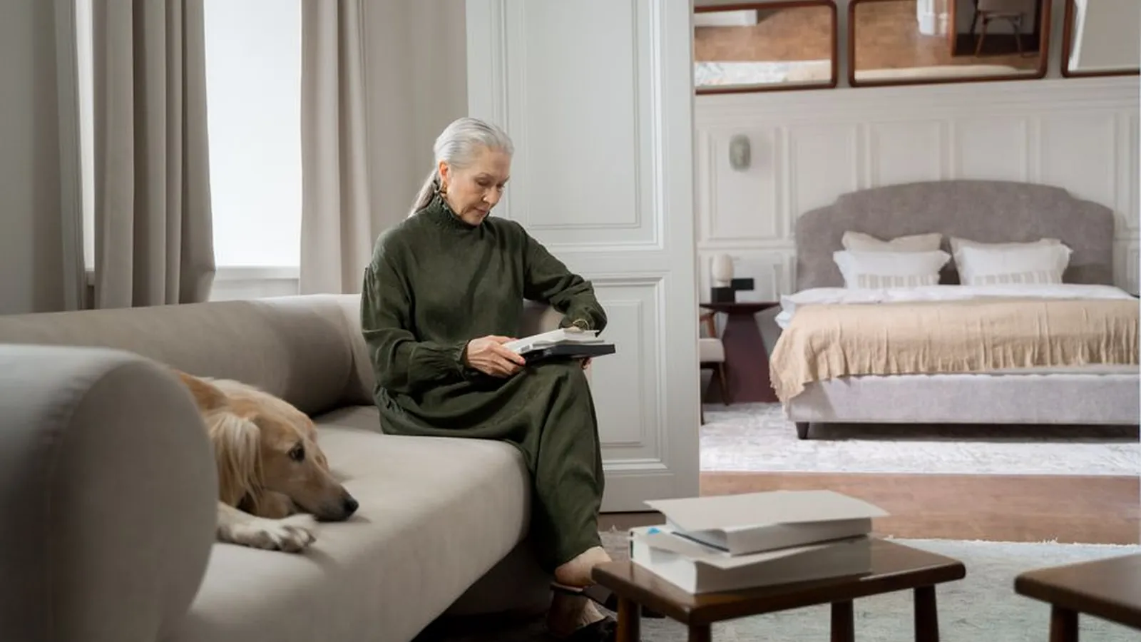 A gray-haired woman sits on a sofa at home reading, while a golden dog rests its head on the cushion beside her.
