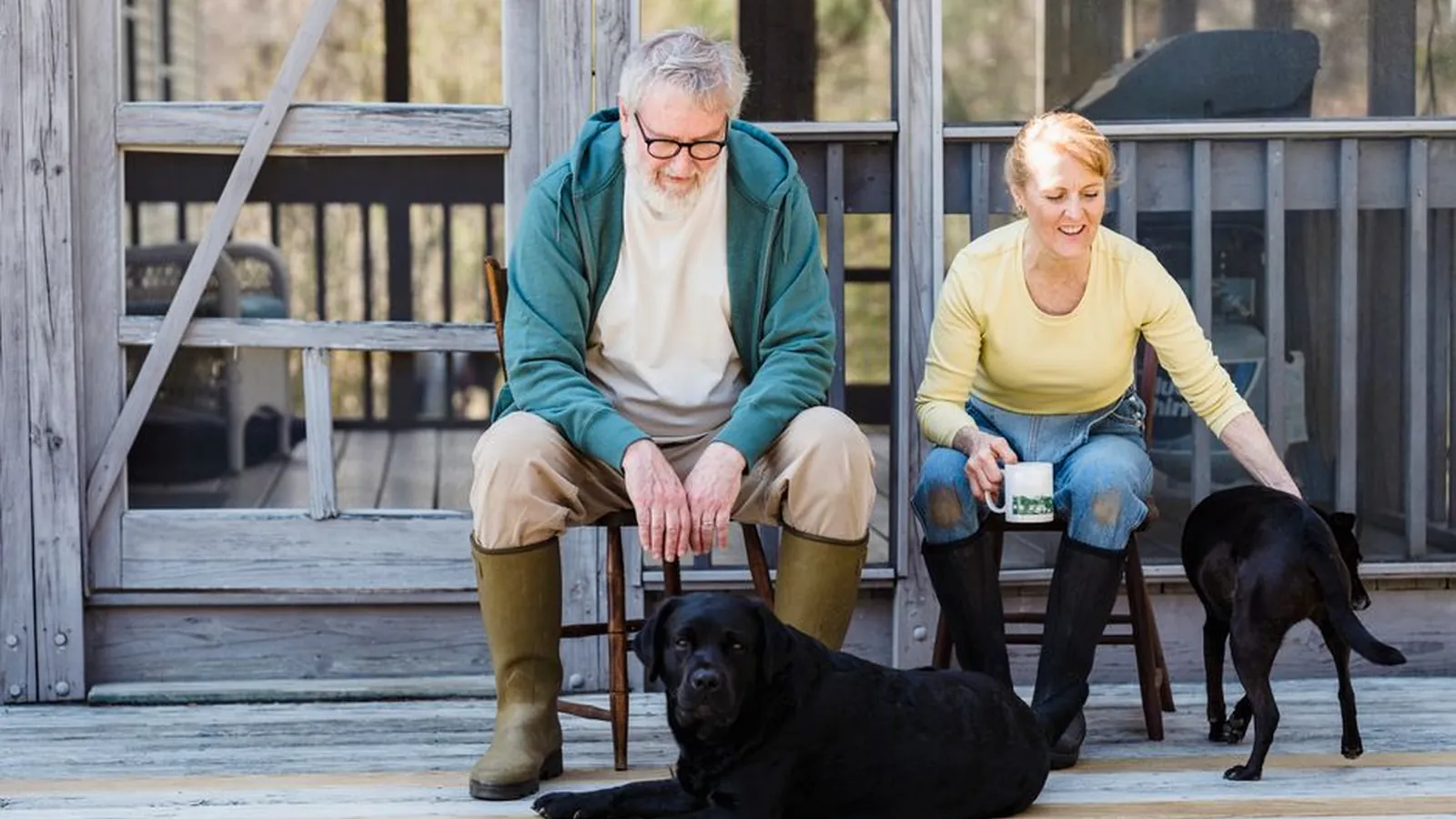 An older man and woman sitting on a wooden porch with a mug of tea, a black Labrador lying at their feet and a second black dog beside them.